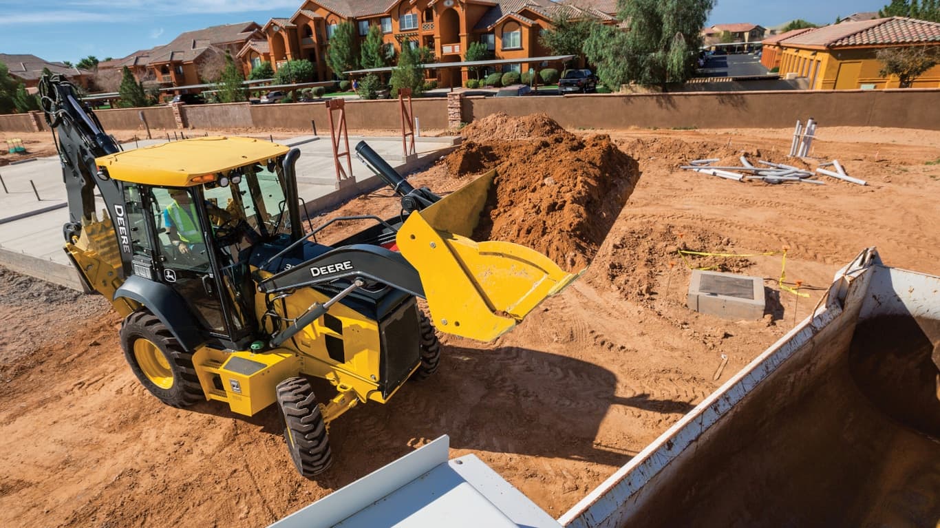A 310G Backhoe moving dirt at a housing worksite with the house foundation in the background. | 4re Equipment | ID: 310GBH