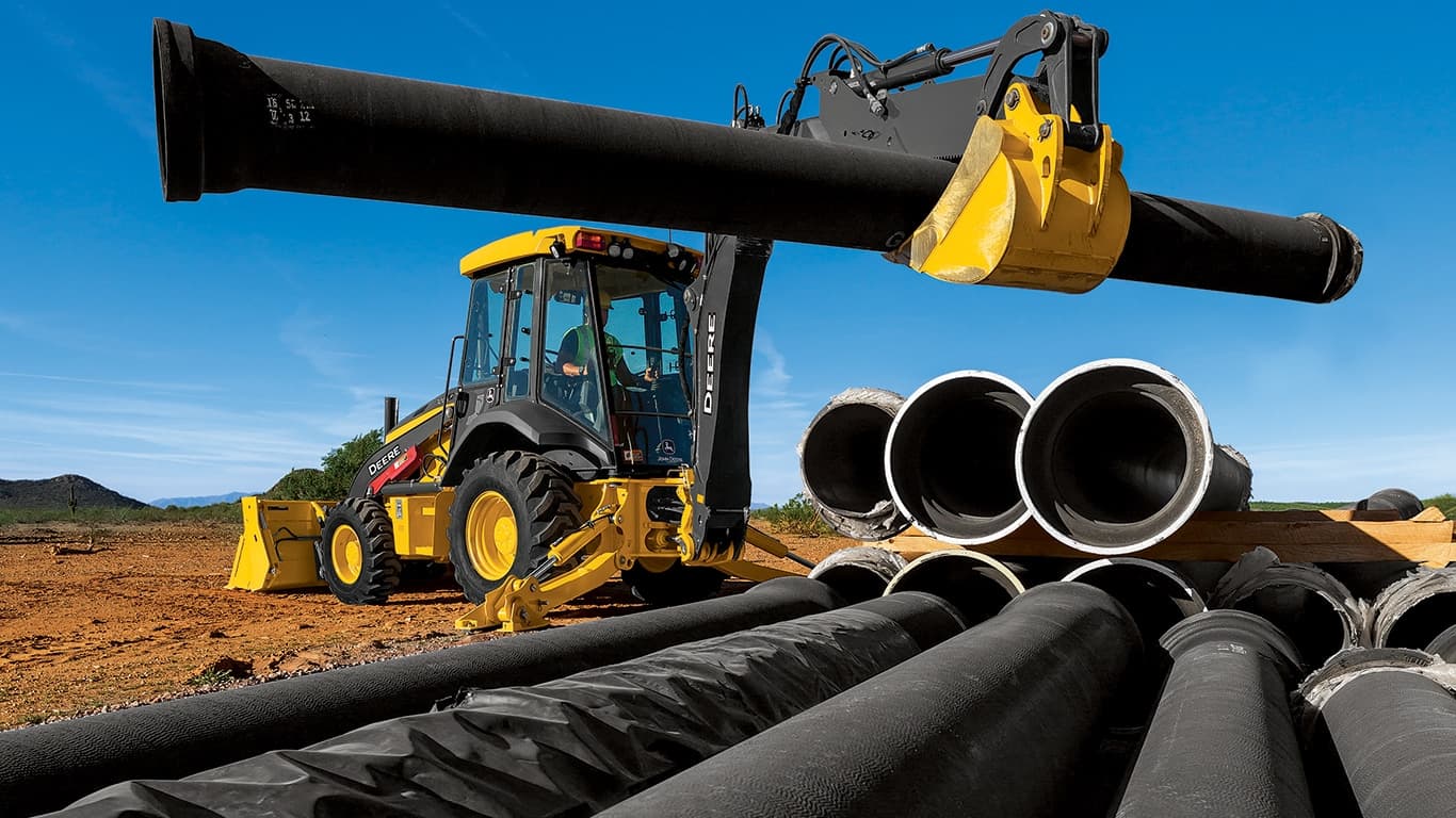 A 310G Backhoe using the boom arm to move large black pipes. | 4re Equipment | ID: 310GBH