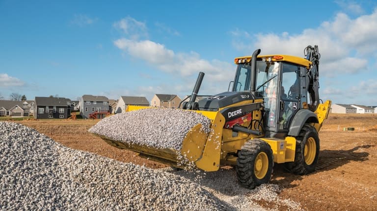 A 320P Backhoe hauling small rocks in the bucket with houses in the background. | 4re Equipment