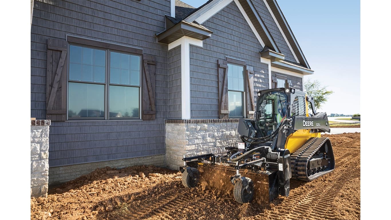 A 325G Compact Track Loader with a power rake attachment raking dirt in front of a house. | 4re Equipment | ID: 325gxt