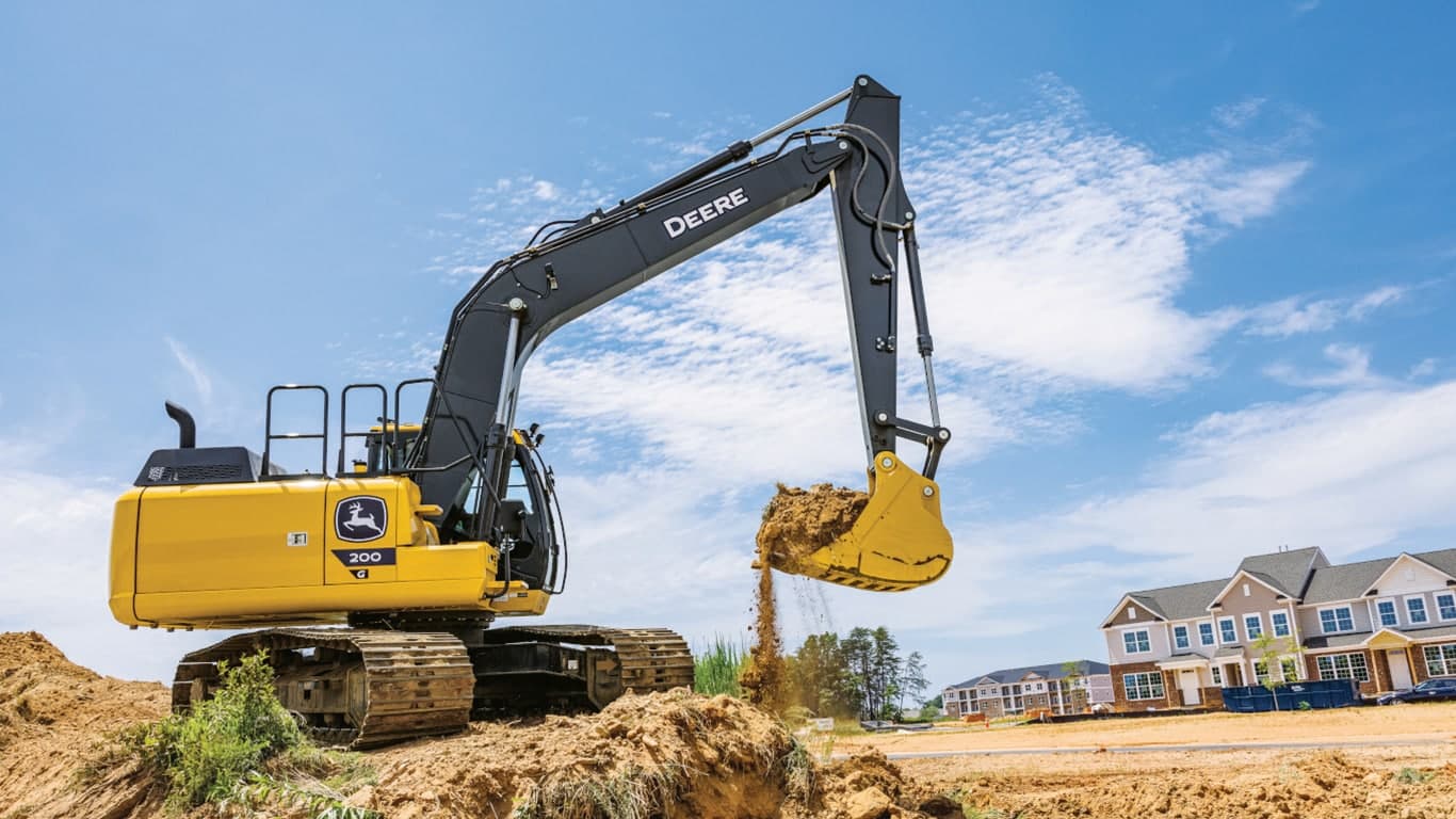 A 200G Excavator moving dirt at a worksite with a housing development in the background. | 4re Equipment | ID: 200GEXFT4