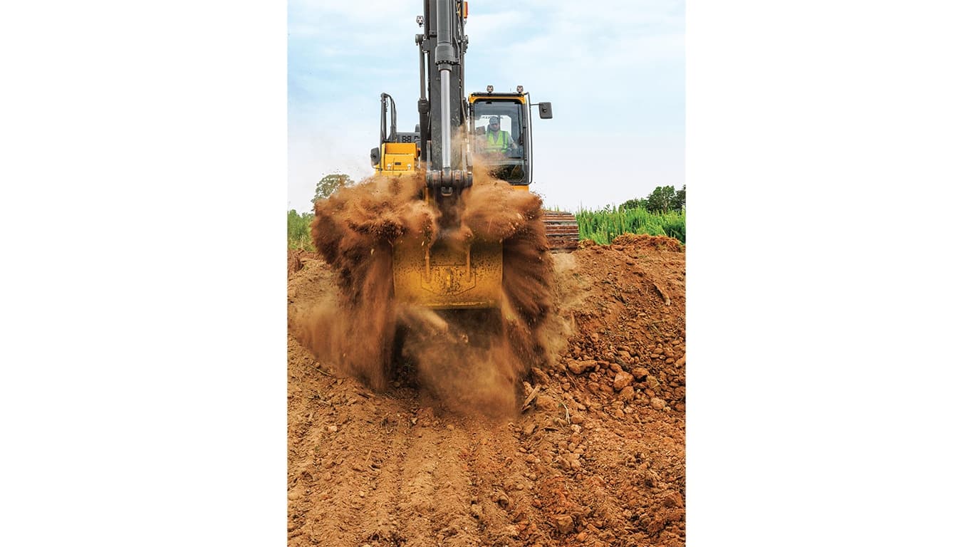 A front-facing perspective of an operator scooping up dirt with a 210P-Tier Excavator with a green field in the background. | 4re Equipment | ID: 210PEXFT4