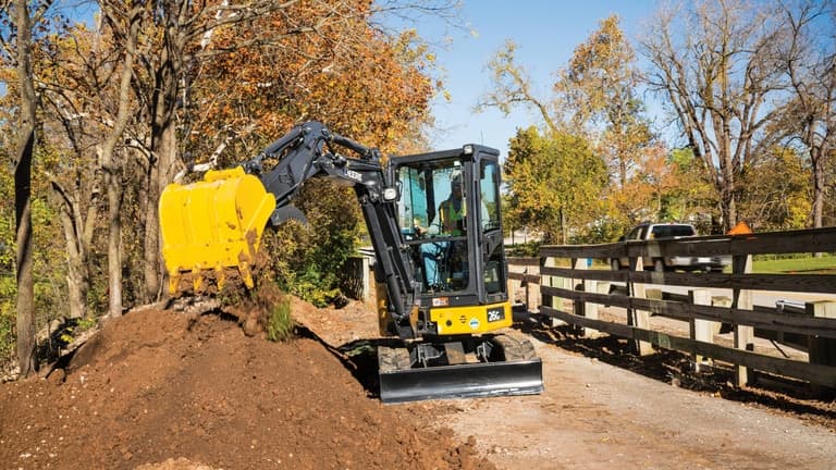 26G Excavator dropping topsoil into a stockpile on a sidewalk in front of a wooden fence. | 4re Equipment