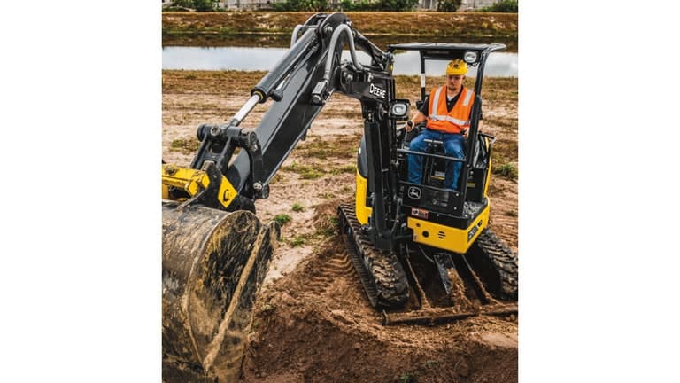 An operator using a 26P-Tier Excavator to scoop dirt at a worksite with a pond in the background. | 4re Equipment