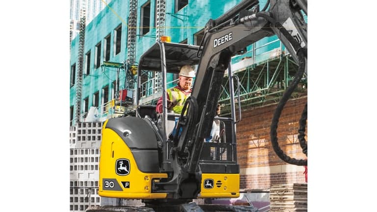 A close-up of an operator using a 30P-Tier Excavator at a worksite with buildings under construction in the background. | 4re Equipment
