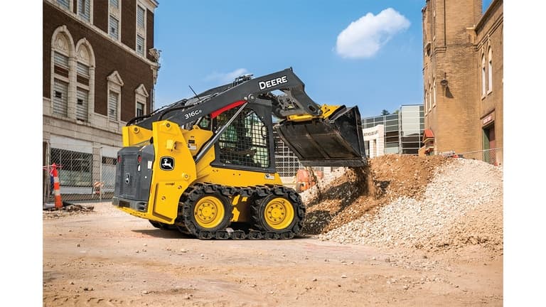 A 316G Skid Steer dumps rocks and dirt into a pile at a worksite with buildings in the background. | 4re Equipment