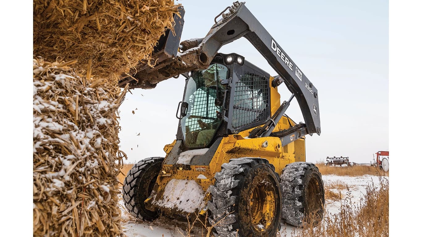 A 330G Skid Steer with bale spears attachment lifting a bale of snowy hay in a snowy field. | 4re Equipment | ID: 330gxt